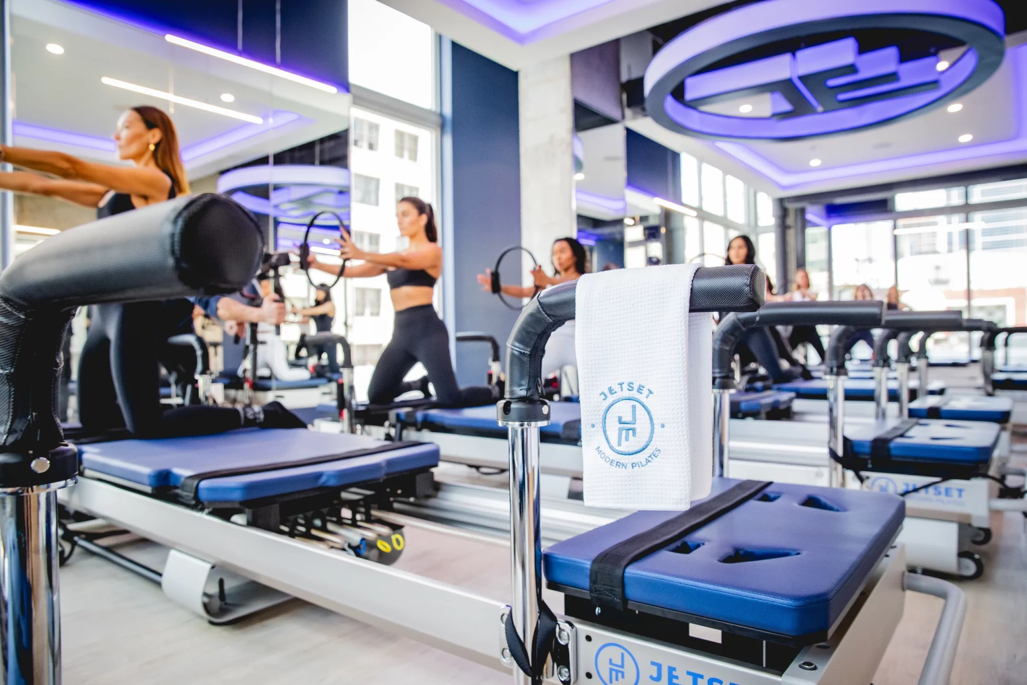 Interior of a JETSET Pilates studio with reformer machines, modern lighting, and clients mid-class in a bright, high-energy space.